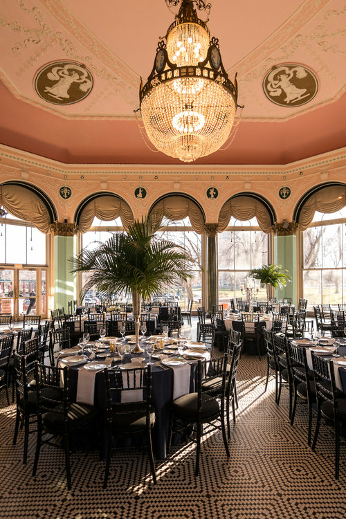 Elegant ballroom set for event with round tables, black chairs, and large crystal chandelier.